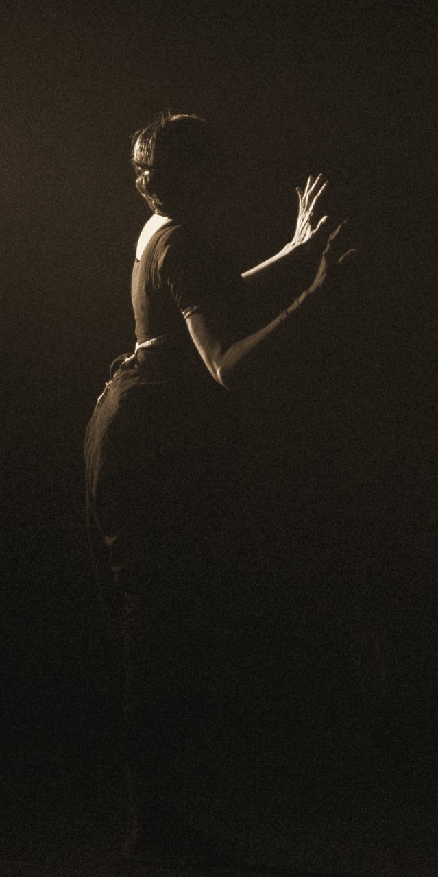 A dancer in soft stage light with raised hands, seen from behind in a hushed, misty theatre